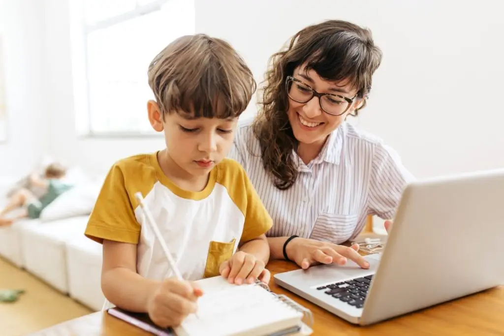 Boy writing on a book while his mother working on laptop. Kid studying with mother working from home.